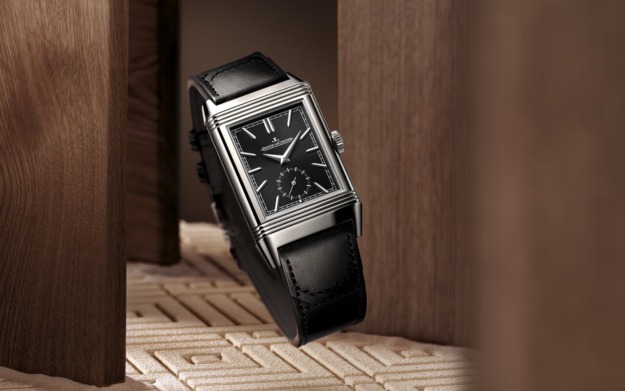 Montre Jaeger-LeCoultre Reverso rectangulaire en acier, cadran noir à petite seconde et bracelet en cuir noir, devant un décor de bois