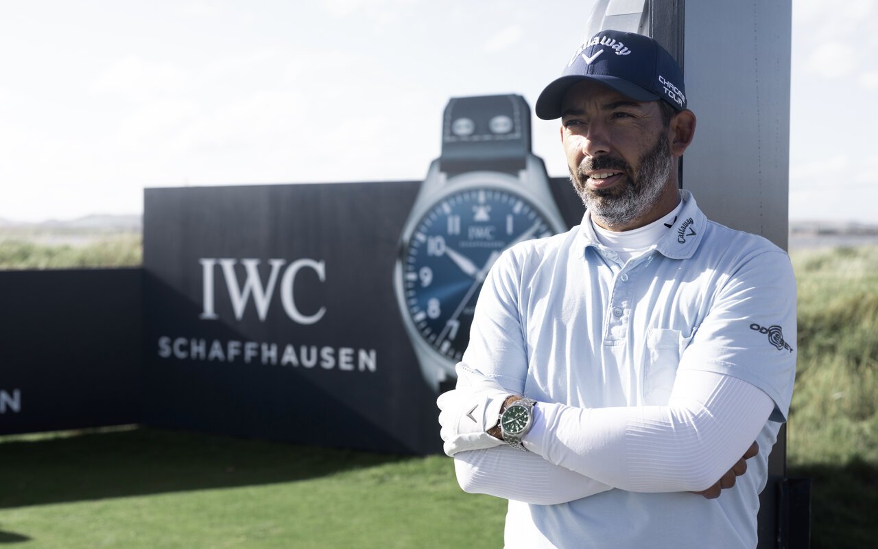 Golfeur en polo bleu clair et casquette marine, bras croisés devant un panneau IWC, une montre chronographe en acier à cadran vert au poignet gauche