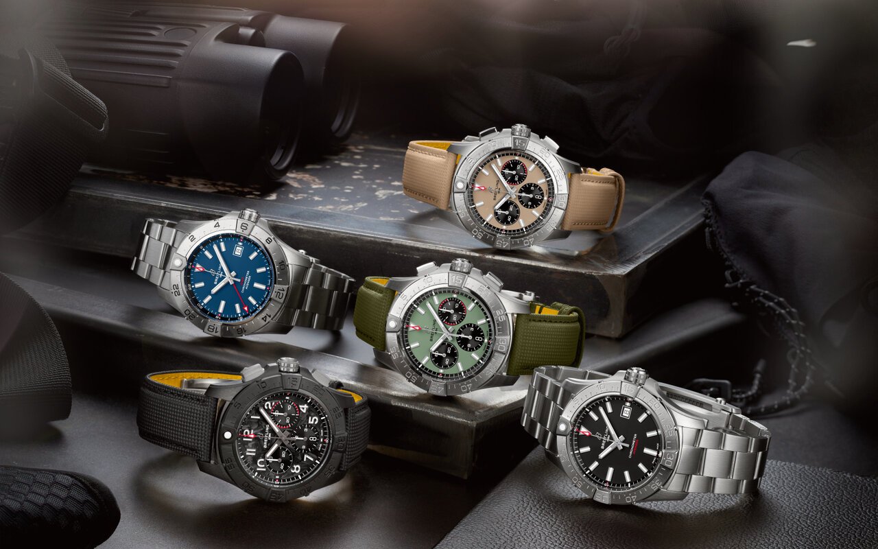 Cinq montres Breitling Avenger alignées sur un établi sombre, cadrans sable, bleu, vert militaire et noir, bracelets acier et textile