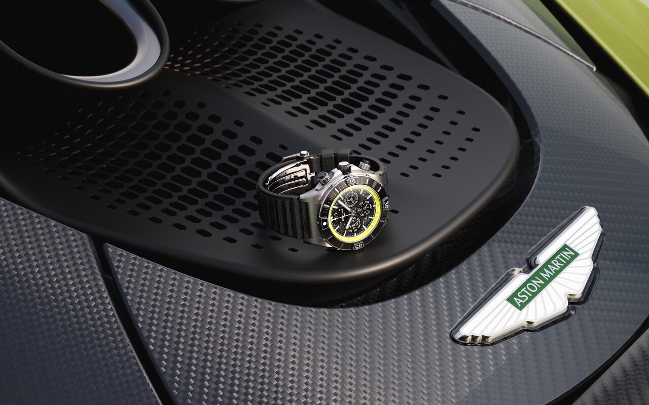 Chronographe Breitling Super Chronomat en titane à cadran noir et rehaut jaune, bracelet caoutchouc noir, posé sur la grille arrière en fibre de carbone d'une Aston Martin Valhalla verte avec l'écusson Aston Martin