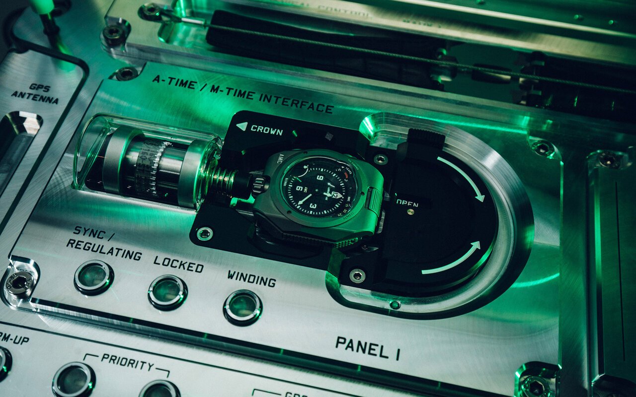 Montre mécanique Urwerk posée dans le berceau de l'horloge atomique AMC, face à un panneau en aluminium portant les commandes de synchronisation, de réglage et de remontage, éclairé en vert